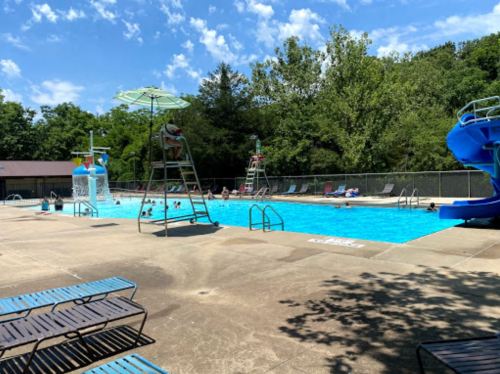 A sunny pool area with a slide, lifeguard tower, and people swimming, surrounded by trees and blue sky.