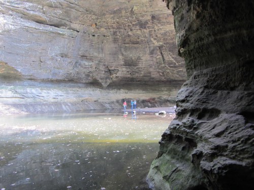 Two people stand in a shallow pool inside a rocky cave, surrounded by textured stone walls and soft lighting.