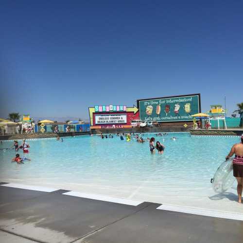 A sunny water park scene with people enjoying the pool and colorful signage in the background.