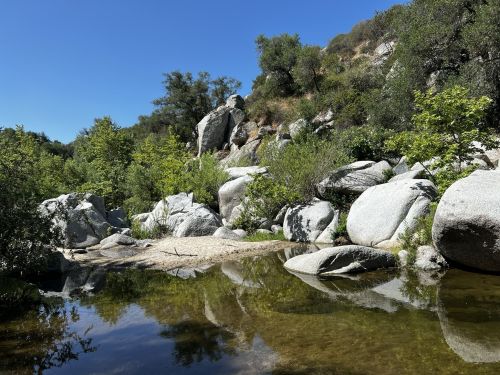 A serene landscape featuring a calm pool of water surrounded by large rocks and lush greenery under a clear blue sky.