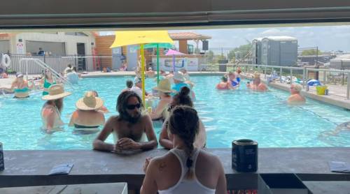 A lively pool scene with people wearing hats, enjoying the water and sun, while others relax at the bar.