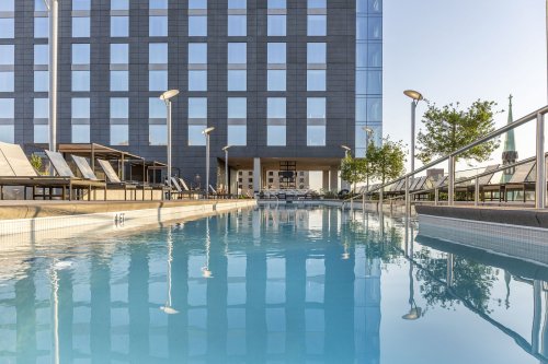 A modern hotel with a reflective pool, lounge chairs, and greenery under a clear blue sky.