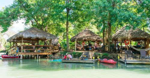 Lush riverside scene with thatched-roof huts, kayaks, and people enjoying the outdoors in a vibrant green setting.