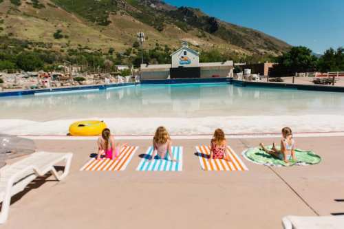 Four children relax on colorful towels by a large, empty swimming pool surrounded by mountains on a sunny day.
