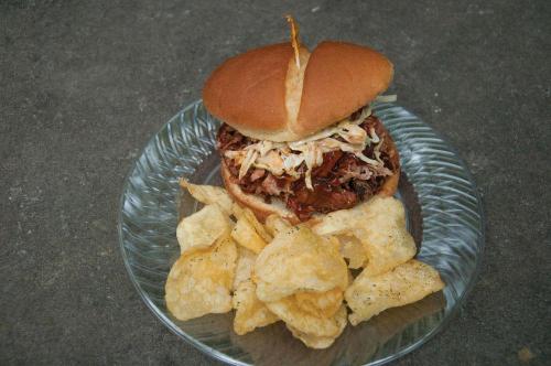 A pulled pork sandwich topped with coleslaw, served on a plate with a side of potato chips.