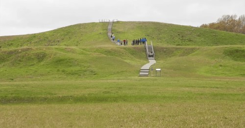 A grassy hill with a staircase leading to the top, where a group of people is walking. Overcast sky in the background.