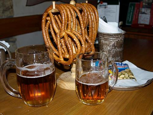 Two mugs of beer sit beside a wooden stand holding twisted pretzels, with snacks in a bowl nearby.