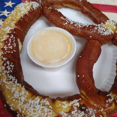 A large pretzel on a plate with a small cup of dipping sauce, set against a background featuring an American flag.
