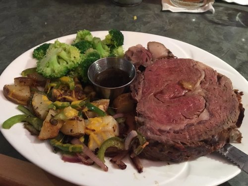 A plate featuring a large slice of prime rib, roasted potatoes with cheese, and steamed broccoli, served with sauce.