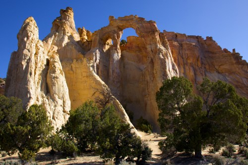 A striking rock formation with an arch, surrounded by green shrubs under a clear blue sky.