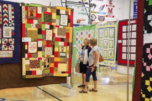 Two women admire colorful quilts displayed at a craft fair in a gymnasium.