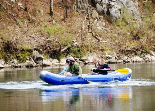 A group of three people paddles a blue inflatable raft on a calm river surrounded by rocky banks and trees.