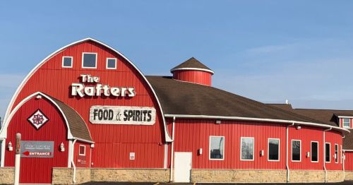 Red barn-style building with a rounded roof, featuring a sign that reads "The Rafters Food & Spirits." Clear blue sky.