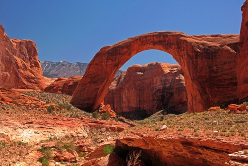 A stunning natural rock arch rises above red sandstone formations under a clear blue sky.