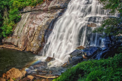 A cascading waterfall flows over rocky cliffs into a serene pool, surrounded by lush greenery and a hint of a rainbow.