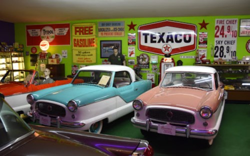 Vintage cars displayed in a colorful retro gas station-themed museum, featuring Texaco and other nostalgic signage.