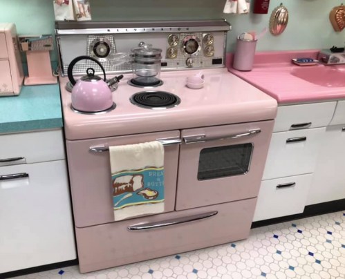 A vintage pink stove with a kettle, towel, and retro kitchen decor in a pastel-colored kitchen setting.