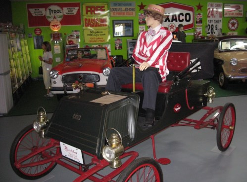 A vintage car display featuring a mannequin in a striped shirt sitting in an old-fashioned vehicle.