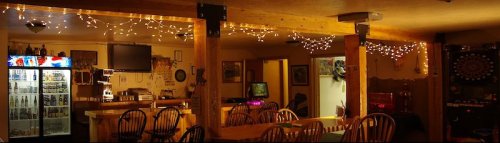 Cozy bar interior with wooden tables, string lights, a fridge, and a dartboard, creating a warm atmosphere.