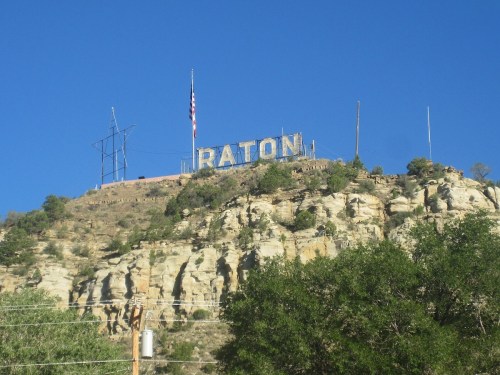 Sign on a hillside reading "RATON," with a clear blue sky and greenery in the foreground.