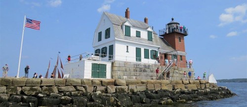A white lighthouse with a red tower on a rocky shore, surrounded by people and an American flag waving nearby.