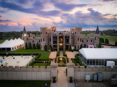 A grand castle with turrets, surrounded by lush greenery, under a colorful sunset sky. Tents are set up in the foreground.