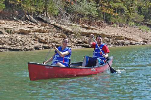 Two people in a red canoe paddling on a calm lake, wearing life jackets and smiling. Trees line the shore in the background.