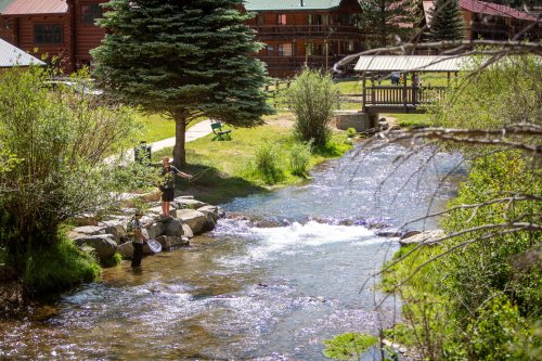 A person fishing by a stream with another nearby, surrounded by greenery and rustic cabins in the background.