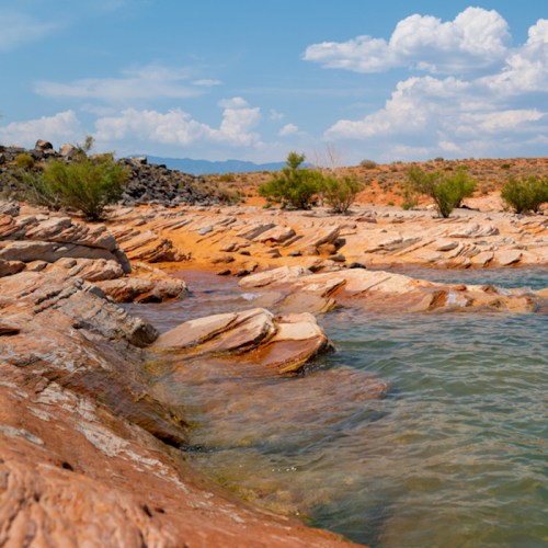 A rocky shoreline with calm water, surrounded by sparse greenery and distant mountains under a partly cloudy sky.