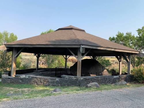 A wooden gazebo with a peaked roof, surrounded by trees and a stone wall, set in a grassy area.