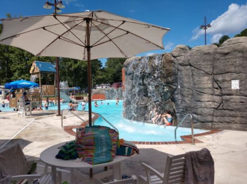 A pool area with a waterfall, umbrellas, and people swimming, surrounded by trees and a playground in the background.