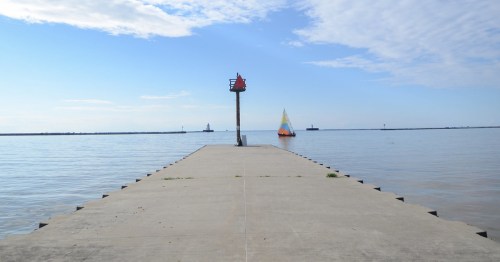 A calm pier extends into the water, with a sailboat in the distance under a clear blue sky.