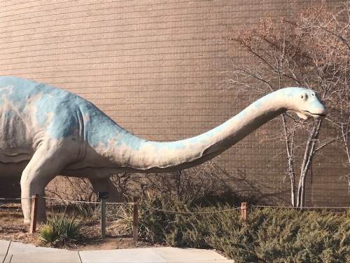 A large, blue dinosaur statue with a long neck stands outside against a brown building and sparse vegetation.