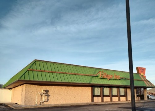 Exterior of a Village Inn restaurant with a green roof and orange signage under a clear blue sky.