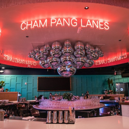 A vibrant bar with a large disco ball and neon sign reading "Cham Pang Lanes," featuring colorful decor and glassware.