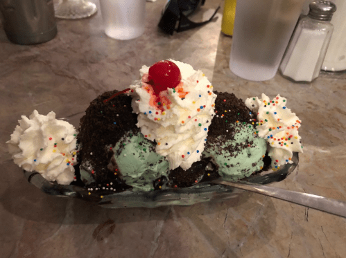 A colorful ice cream sundae with mint chocolate chip ice cream, whipped cream, chocolate crumbs, and a cherry on top.