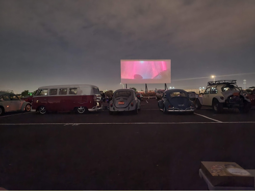 A drive-in movie scene with vintage cars parked in front of a large screen displaying a colorful film under a night sky.