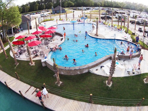 Aerial view of a lively pool area with people swimming, surrounded by palm trees and colorful umbrellas.