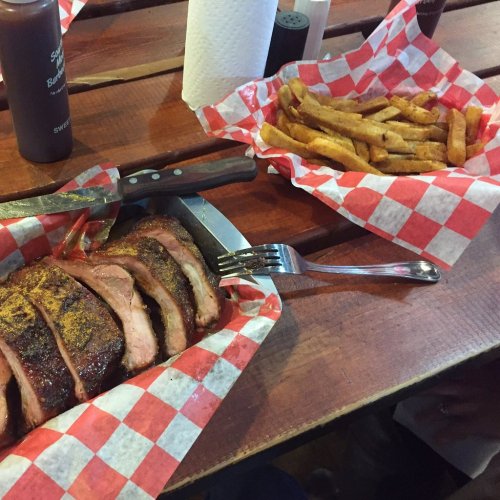 A plate of sliced smoked meat with a fork, accompanied by a basket of golden fries on a wooden table.