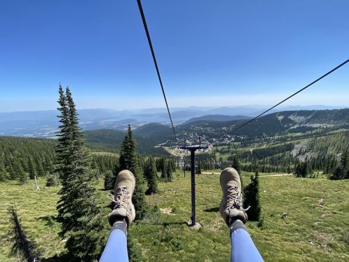 View from a chairlift with feet dangling above a green landscape and distant mountains under a clear blue sky.