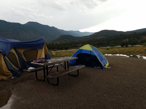 A campsite with two tents and a picnic table, surrounded by mountains and a cloudy sky.