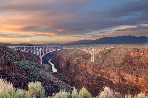 A stunning bridge spans a deep canyon at sunset, with mountains in the background and a river below.