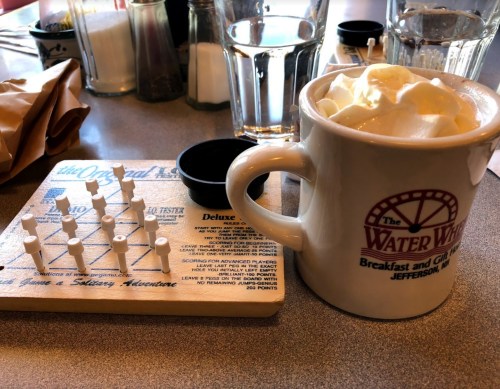 A mug of whipped cream-topped beverage next to a peg game on a diner table.