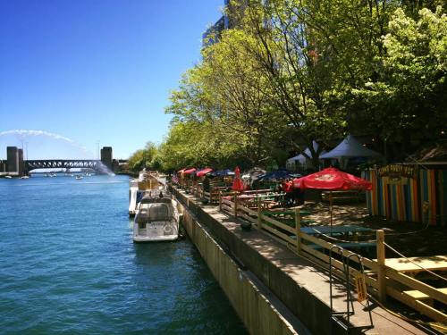 A sunny riverside scene with colorful umbrellas, boats, and trees lining the water's edge.