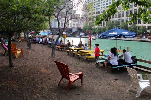 Outdoor seating area by a river, with colorful tables, umbrellas, and people enjoying the scenery.