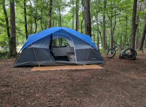 A blue and gray tent set up in a wooded area, with a bicycle parked nearby on the forest floor.