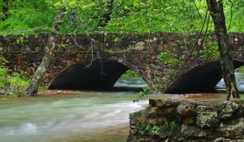 A stone bridge with two arches spans a flowing river, surrounded by lush green trees and foliage.