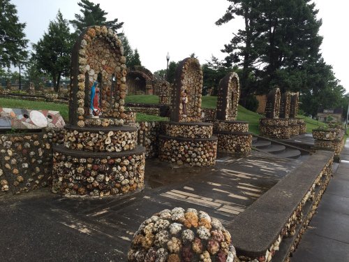 A unique outdoor shrine adorned with colorful shells and stones, featuring arches and a statue, surrounded by greenery.