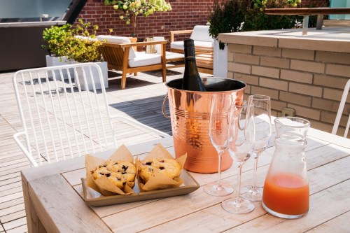 A sunny outdoor setting with a table featuring muffins, a wine bucket, glasses, and a pitcher of juice.