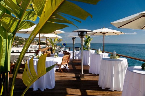 A scenic outdoor terrace with tables, umbrellas, and ocean views, surrounded by tropical plants.